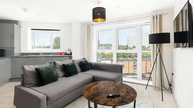 A modern living room with a grey sofa, a round coffee table, a floor lamp, and a large window with a city view. - St Lukes Retreat | Modern Top Floor City Escape (West Midlands)