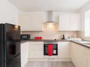 A modern kitchen with a black refrigerator, a microwave, a stove, and a red oven mitt hanging on the oven handle. - Birmingham Estate | Gather Stay & Celebrate (Winson Green)
