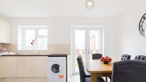 A modern kitchen with a built-in washing machine, a dining area with a table and chairs, and a large wall clock. - Birmingham Estate | Gather Stay & Celebrate (Winson Green)