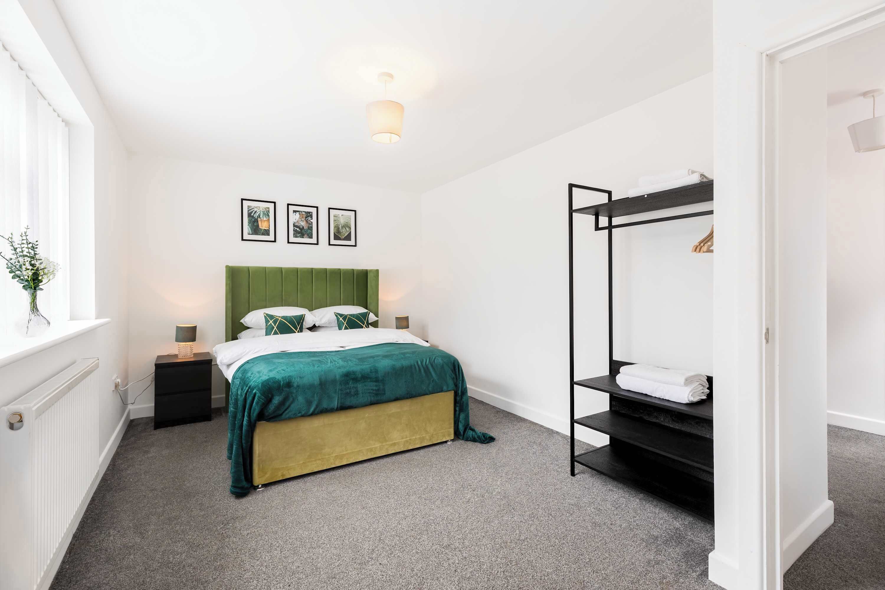 Ein Schlafzimmer mit einem Bett, Nachttischen, einer grünen Kopfteil, einem schwarzen Metallregal und einem Fenster mit einer Pflanze.