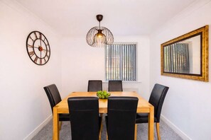 A dining room with a wooden table, black chairs, a fruit bowl, a wall clock, a pendant light, a window with blinds, and a framed picture. - Anchor House | Spacious Group Stay | Free Parking (West Midlands)