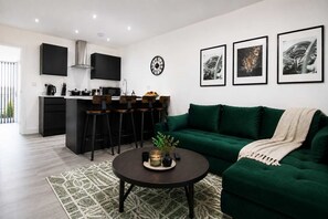 A modern living room with a green sofa, a coffee table, and a kitchen area with a bar stool set. - Cardiff Bloc 2 | Couple & Groups | Large Balcony (Rumney)