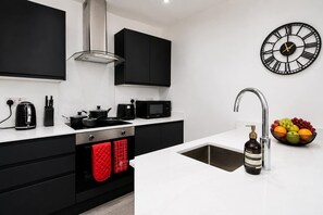 A modern kitchen with black cabinets, a white countertop, a stainless steel sink, a black microwave, a gas stove, and a wall clock. - Cardiff Bloc 2 | Couple & Groups | Large Balcony (Rumney)