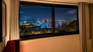 Ett rum med ett stort fönster med utsikt över en stadssiluett i natt, där upplysta byggnader och en bro syns.