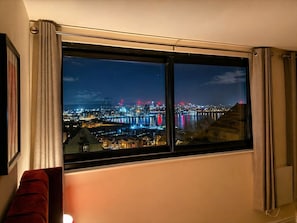 Ein Zimmer mit großem Fenster mit Blick auf eine beleuchtete Stadtlandschaft bei Nacht, darunter Gebäude und eine Brücke.