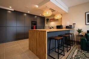 Moderni keittiö, jossa on puinen keittiösaareke, baarituolit, riippuvalaisimet ja vihreä sohva.