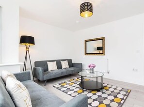 Een moderne woonkamer met een grijze bank, een ronde salontafel, een kamerkleed met dessin, een vloerlamp en een hanglamp.