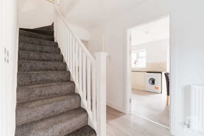 A staircase with a white railing leading to a kitchen area with a washing machine. - Birmingham Estate | Private Gated Group Escape (Winson Green)