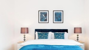 Une chambre d’hôtel avec un lit, deux œuvres d’art encadrées, une table de chevet avec une lampe et une petite table à côté du lit.