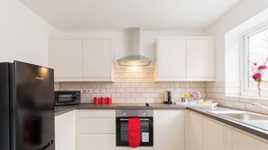 A modern kitchen with a black refrigerator, a microwave, a stove, and a red oven mitt hanging on the oven handle. - Birmingham Estate | Gated | Built For Large Groups (Winson Green)