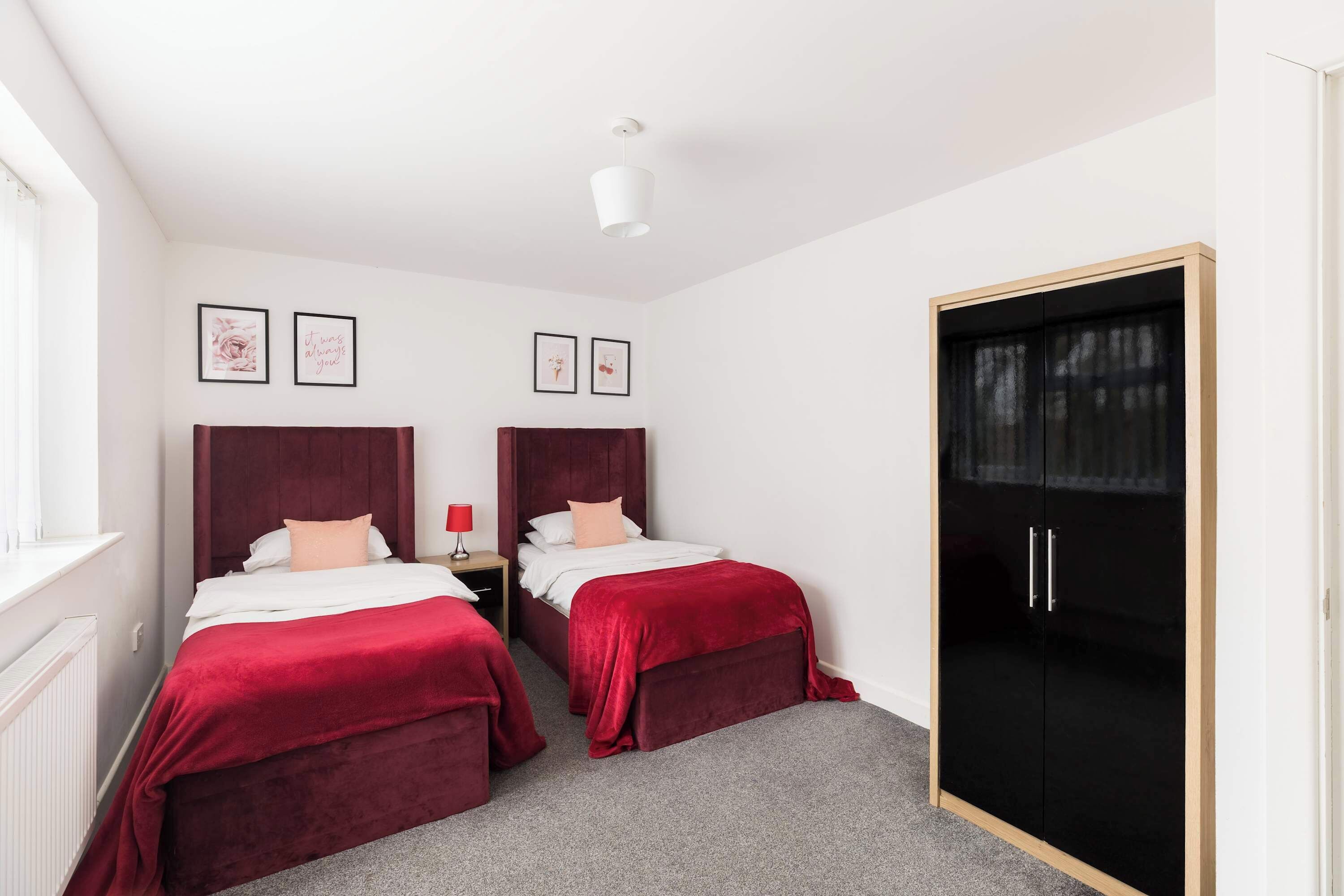 Quarto de hotel com duas camas, um guarda-roupa, uma mesinha de cabeceira e quadros decorativos na parede.