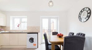 A modern kitchen with a built-in washing machine, a dining area with a table and chairs, and a large wall clock. - Birmingham Estate | Gated | Built For Large Groups (Winson Green)