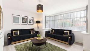 Een woonkamer met twee zwarte banken met gouden accenten, een ronde salontafel, een vloerlamp en een groot raam met zonwering.