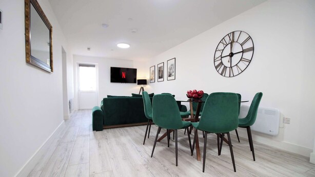 Un salon moderne avec un canapé, des fauteuils verts, une table à manger, un téléviseur et une horloge murale.