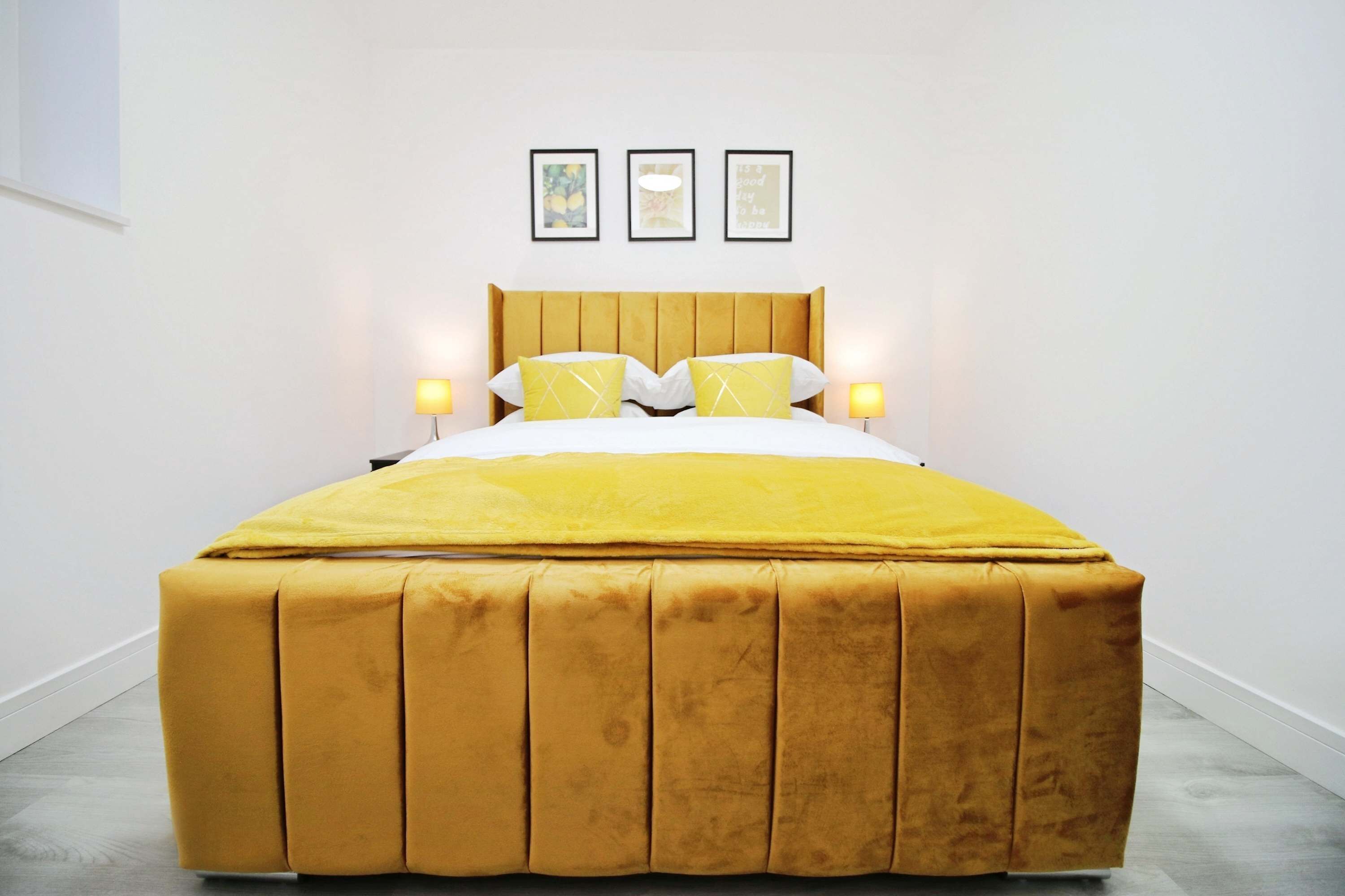 A bed with a yellow headboard, two bedside lamps, and three framed pictures on the wall.