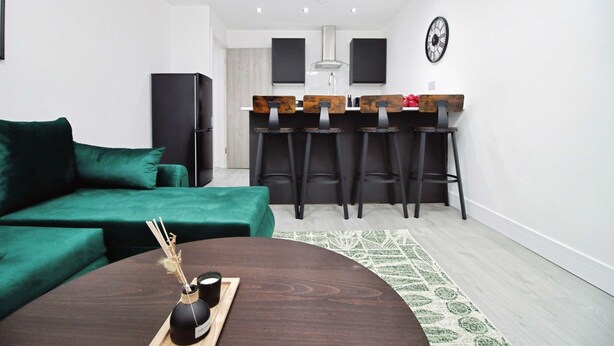 Un salon moderne avec un canapé vert, une table basse en bois et un coin cuisine équipé de tabourets de bar.