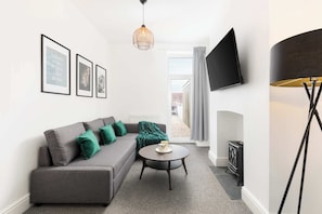 Ein modernes Wohnzimmer mit grauem Sofa, grünen Kissen, einem runden Couchtisch, einer Pendelleuchte, einem Wand-Fernseher und einem Kamin.