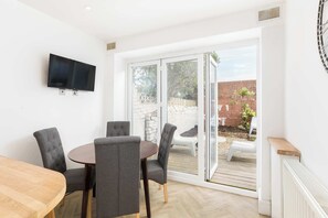 Ein Zimmer mit einem runden Tisch, vier grauen Polsterstühlen, einem an der Wand montierten Flachbildfernseher und einer Glastür, die zu einem Außenbereich mit einer Ziegelwand und Pflanzen führt.