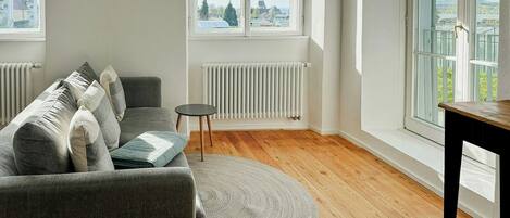 Un salon avec un canapé gris, un tapis rond, une petite table et de grandes fenêtres donnant sur des immeubles et des arbres.