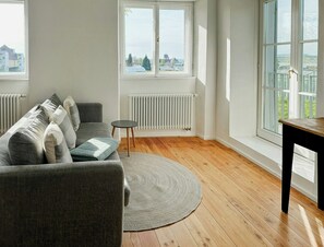 Ein Wohnzimmer mit grauem Sofa, einem runden Teppich, einem kleinen Tisch und großen Fenstern mit Blick auf Gebäude und Bäume.