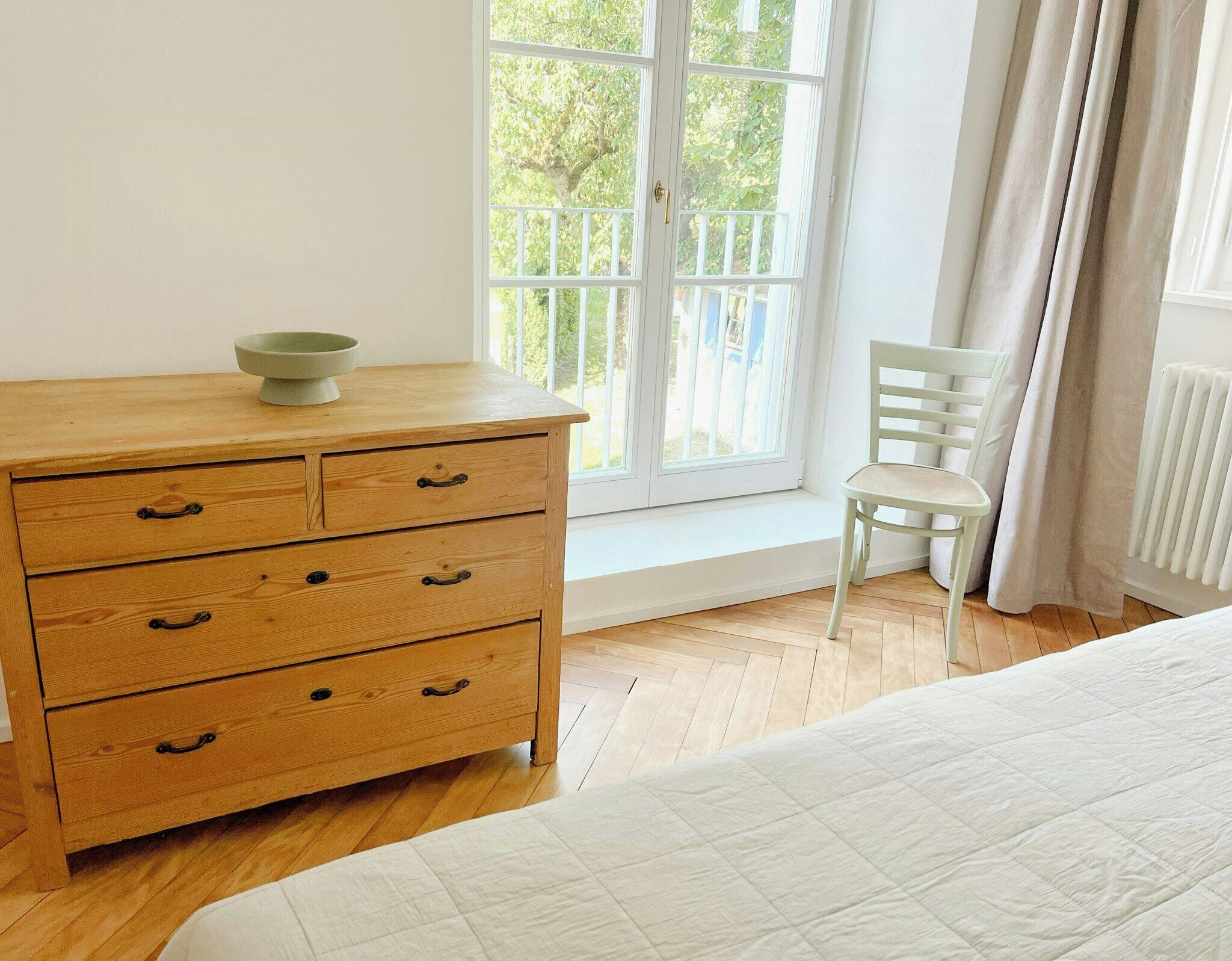 Une chambre à coucher avec une commode en bois, une chaise, un lit, une fenêtre avec des rideaux et un radiateur.