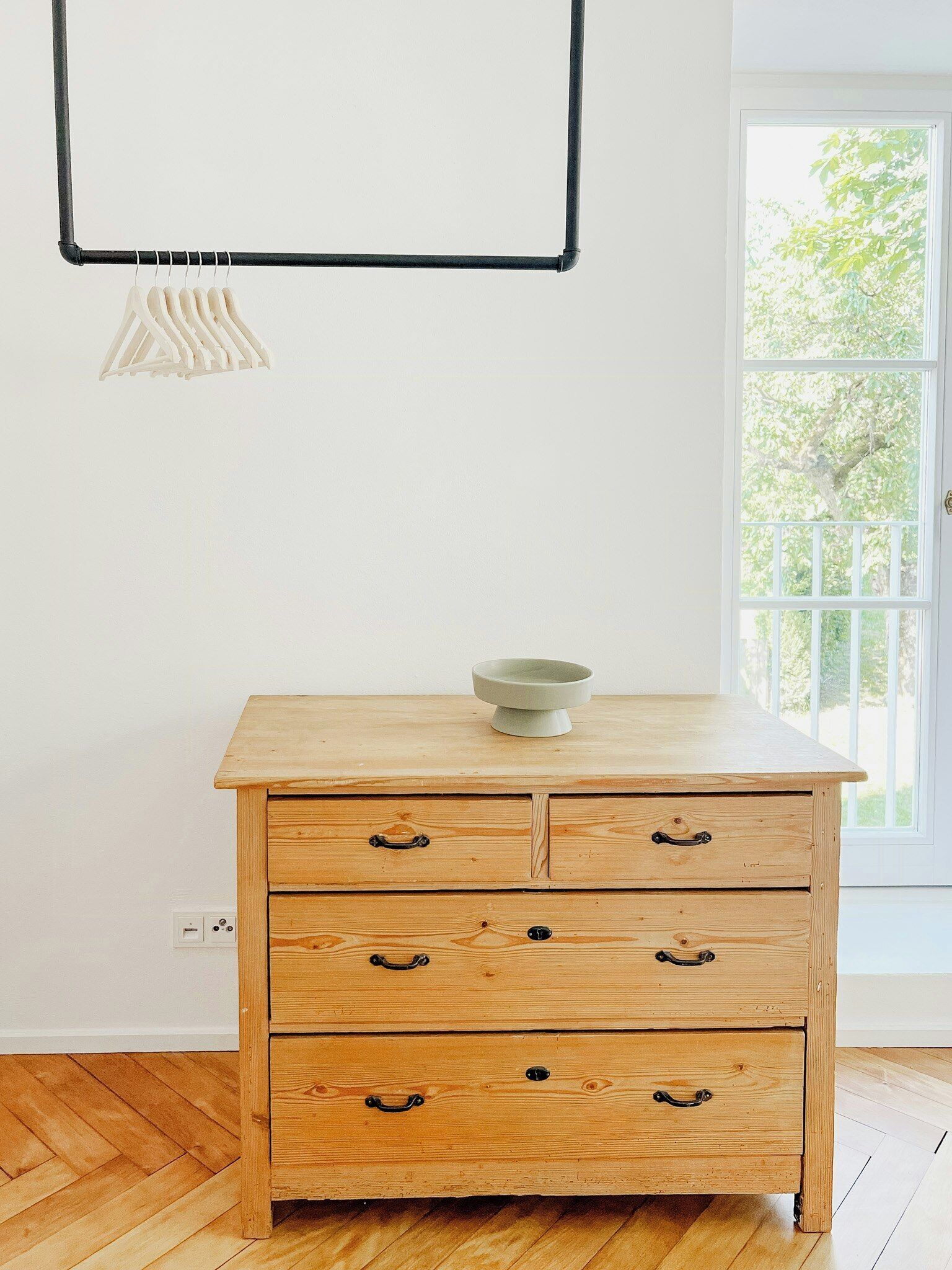 A wooden dresser with drawers and a bowl on top, a hanging light fixture, and a window with a view of trees.