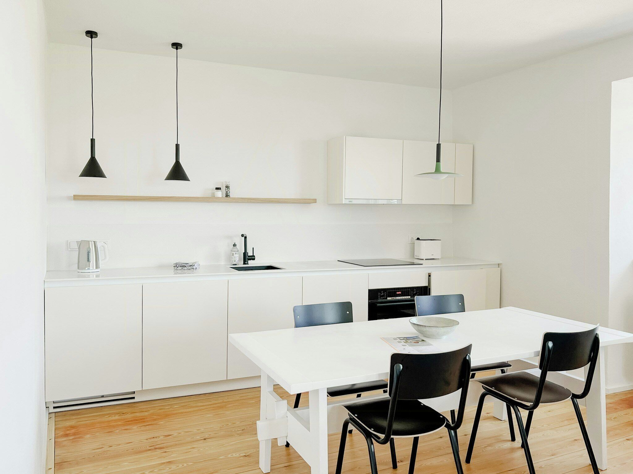 A modern kitchen with white cabinets, a white countertop, two pendant lights, a kettle, a toaster, and a dining table with four chairs.