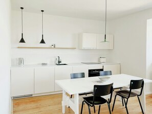 Eine moderne Küche mit weißen Schränken, einer weißen Arbeitsplatte, zwei Pendelleuchten, einem Wasserkocher, einem Toaster und einem Esstisch mit vier Stühlen.
