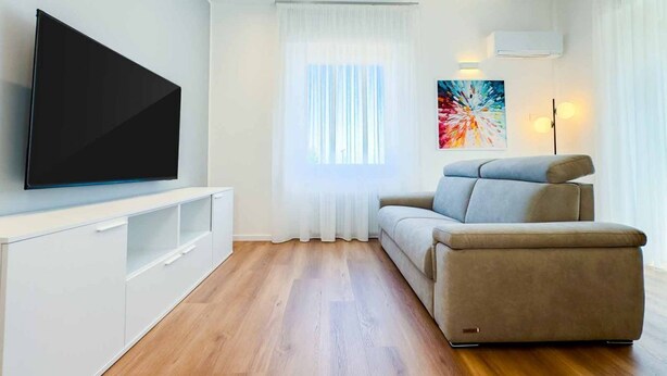 Una sala de estar moderna con una televisión de pantalla plana, un sofá, un mueble blanco para entretenimiento y un ventanal con cortinas translúcidas.