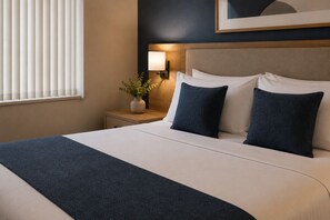Ein ordentlich gemachtes Bett mit einem dunkelblauen Überwurf, ein Nachttisch mit Blumenvase und eine Wandlampe.