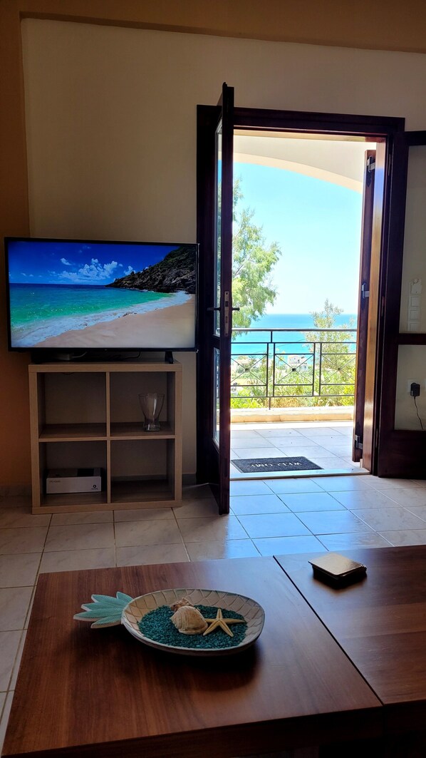 A living room with a television displaying a beach scene, a wooden table with decorative items, and a view of the sea through an open door.