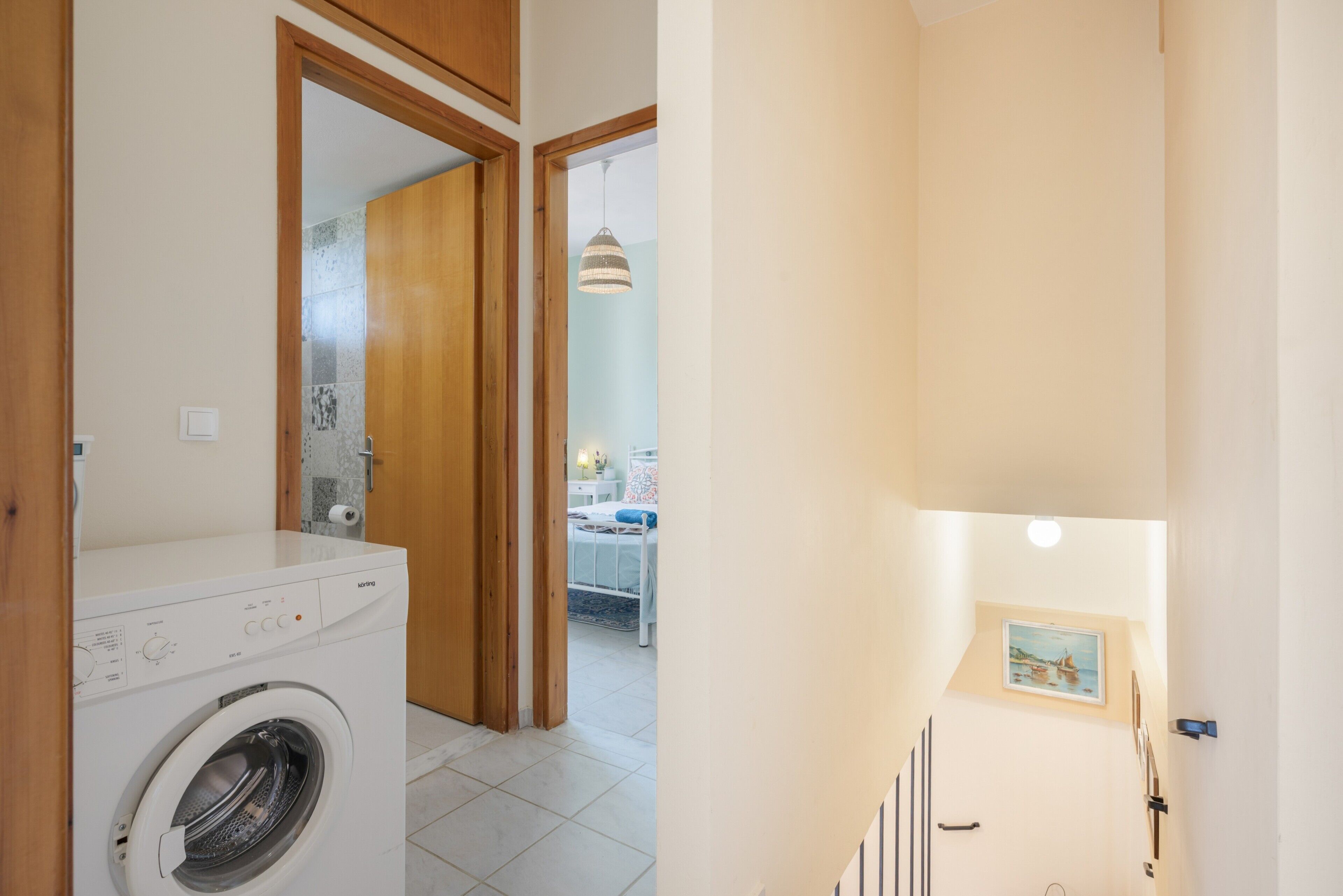 A laundry room with a washing machine, a wooden door, and a view into a bedroom with a bed and a hanging light fixture.