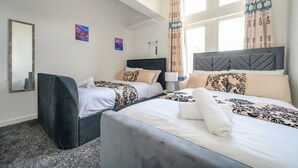 Habitació d'hotel amb dos llits, una finestra gran amb cortines, un mirall i una taula petita amb una làmpada.