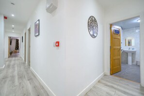 Een gang met een houten vloer, een brandmelder, een ingelijst kunstwerk, een decoratief object aan de muur, en een badkamer zichtbaar door een open deur.