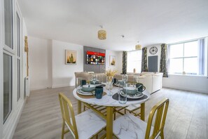 Un coin repas avec une table ronde dressée pour quatre personnes, un canapé, une bibliothèque et une grande fenêtre avec des rideaux.