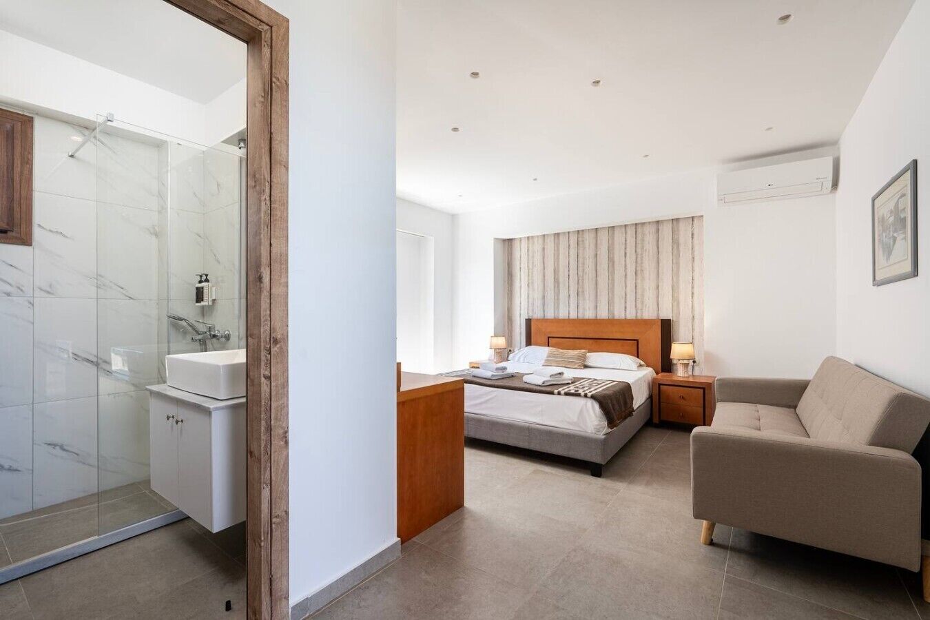 A modern hotel room with a glass shower enclosure, a bed with a wooden headboard, a bedside table with lamps, and a beige armchair.