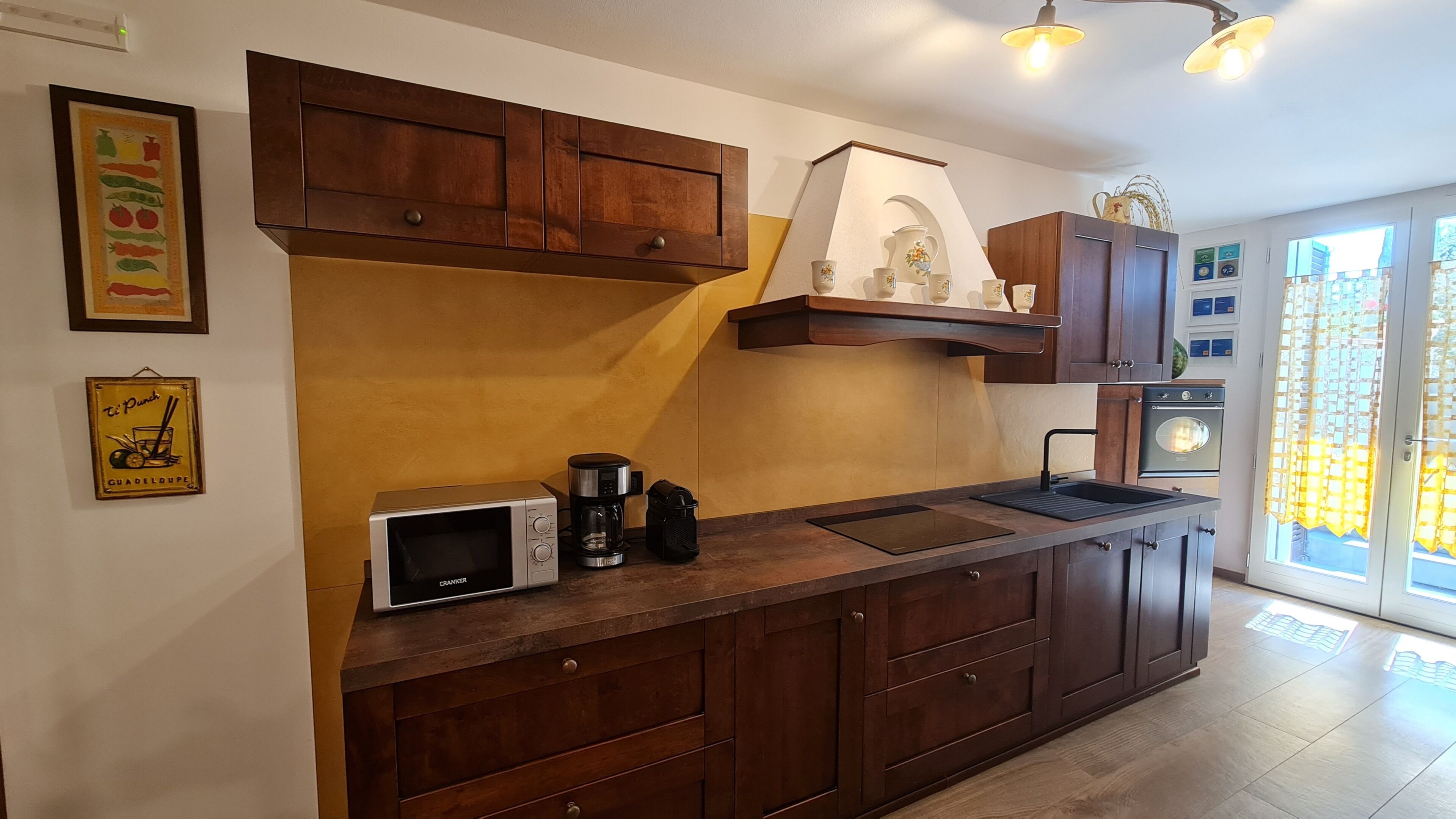 Una cocina con armarios de madera, microondas, cafetera y ventana con cortinas.