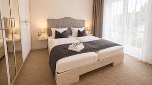 Ein Doppelbett mit gebogenem Polsterkopfteil, Nachttische mit Lampen, ein großes Fenster mit durchsichtigen Vorhängen und ein Kleiderschrank mit Spiegelüren.
