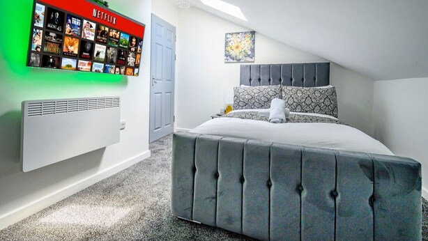 A modern bedroom with a grey tufted bed, a wall-mounted Netflix display, and a painting above the bed. - Stylish Home for 6 in West Yorkshire (West Yorkshire)