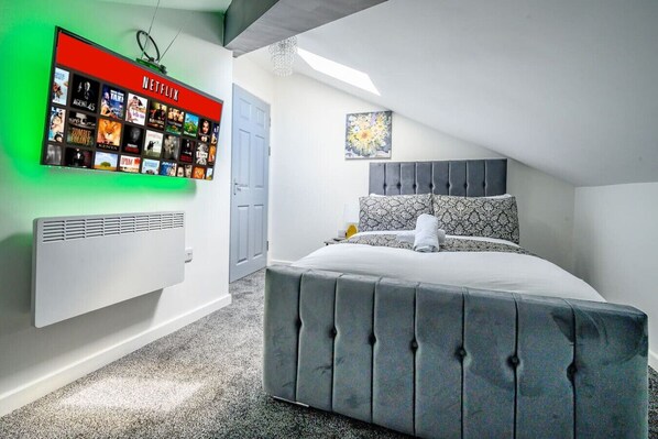 Une chambre à coucher moderne avec un lit capitonné gris, un écran Netflix fixé au mur et un tableau au-dessus du lit.