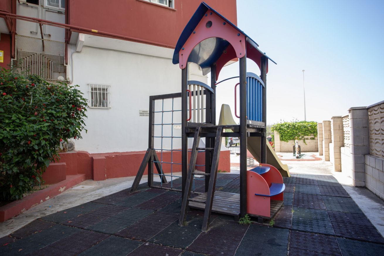 A children's play structure with a slide and climbing area, adjacent to a building with a red exterior and a gated entrance.