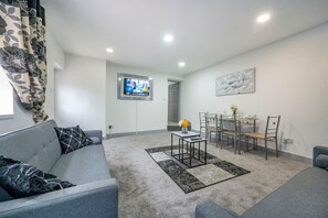 Ein modernes Wohnzimmer mit einem grauen Sofa, einem Esstisch mit Stühlen, einem flachbildfernseher an der Wand und einem Gemälde über dem Tisch.