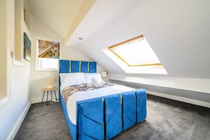 Een slaapkamer met een schuine kap, dakraam, een bed met blauw hoofdeinde en een klein nachtkastje.