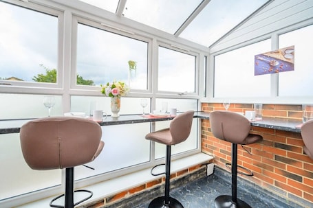 A conservatory with a brick wall, a glass table, and three brown chairs.. H17A Cosy & Welcoming home