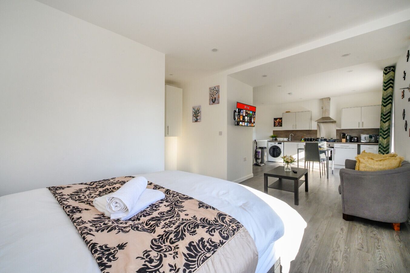 Une chambre d’hôtel moderne avec un grand lit, un petit bureau, un canapé gris et, au fond, une kitchenette.