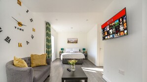 Moderni hotellihuone, jossa on harmaa sohva, valkoisella päällisellä varustettu sänky, musta sohvapöytä ja seinälle asennettu televisio, jonka näytöllä näkyy Netflixin käyttöliittymä.