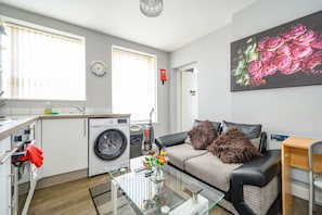 Um apartamento moderno com cozinha, um sofá com almofadas fofas, uma mesa de centro de vidro e um grande quadro com flores na parede.