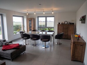 A modern living room with a dining area, large windows, and a view of the outdoors. - Landglück – Vacation home with a terrace, garden, and space for up to 9 guests (Oesterdeichstrich)