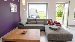 Un salon moderne avec un canapé d’angle gris, une table basse en bois et une grande porte-fenêtre coulissante donnant sur un espace extérieur.