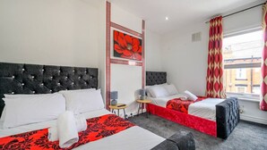 A hotel room with two beds, a decorative wall panel, a small table with a lamp, and a window with curtains.
